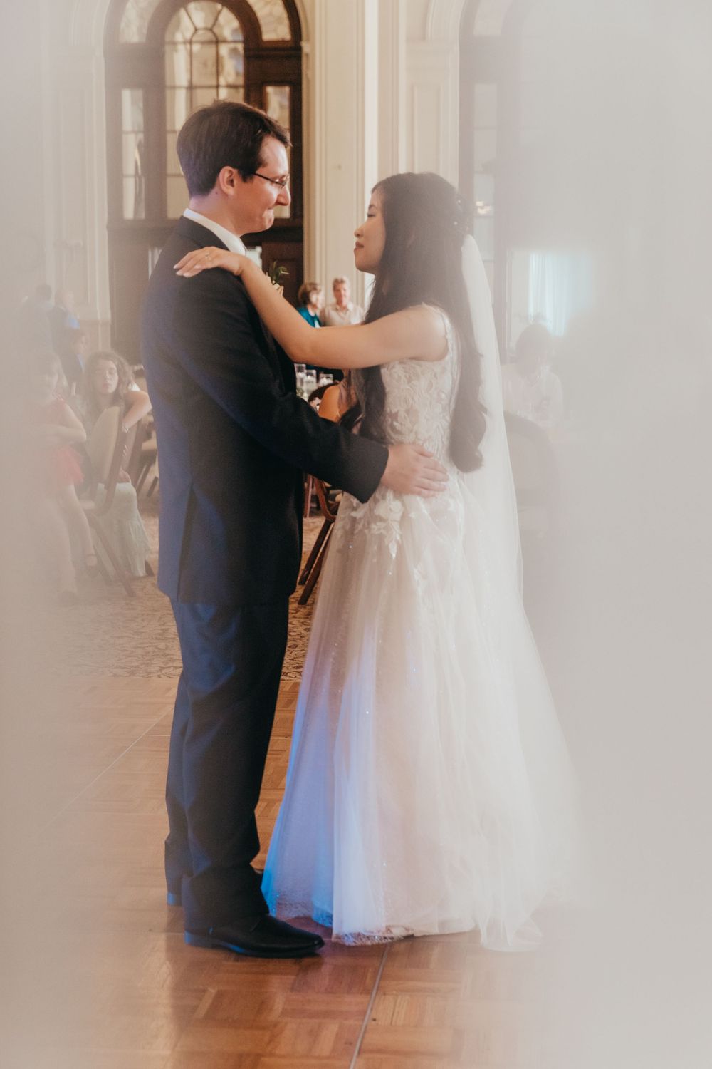 Wedding couple shares a tender dance moment in a sunlit ballroom with arched windows and wooden floor.