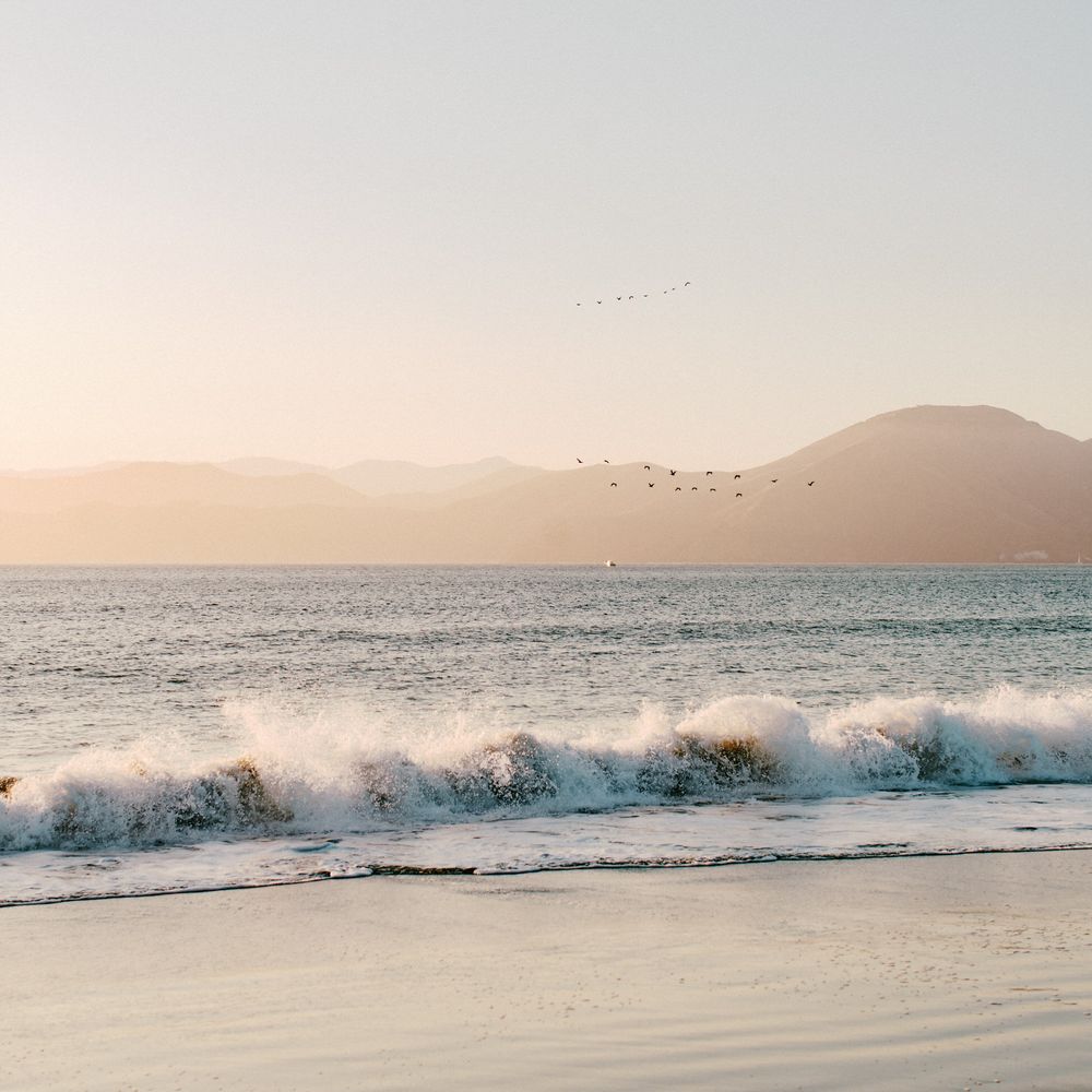Waves crash on a beach at sunset with birds flying over mountains in the distance.
