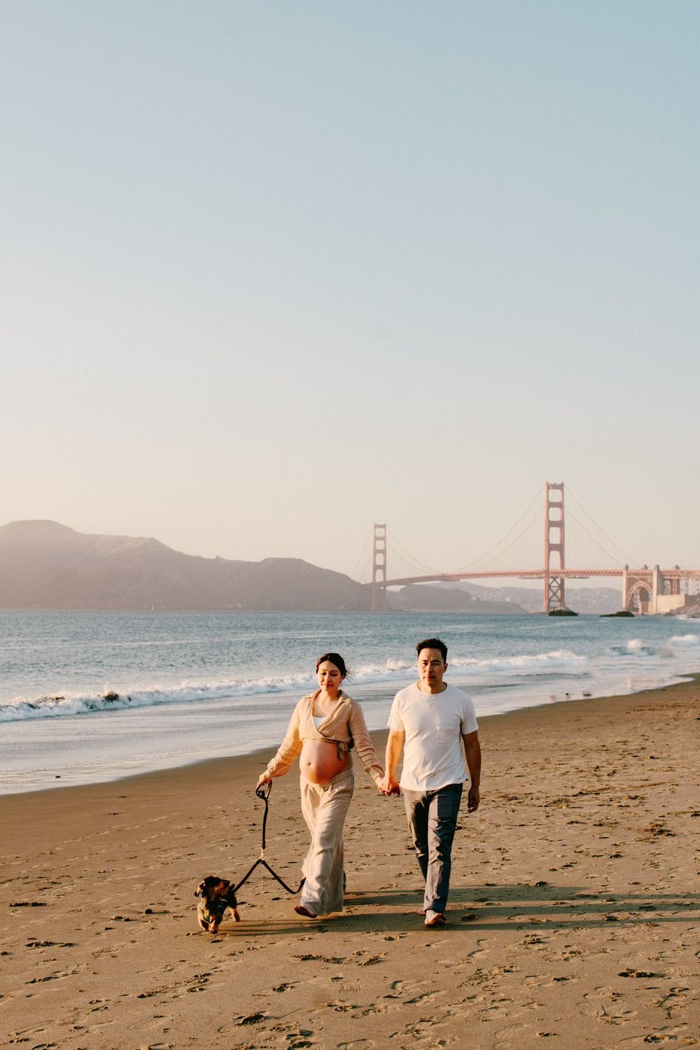 Couple strolling with their small dog along Baker Beach with the Golden Gate Bridge visible.