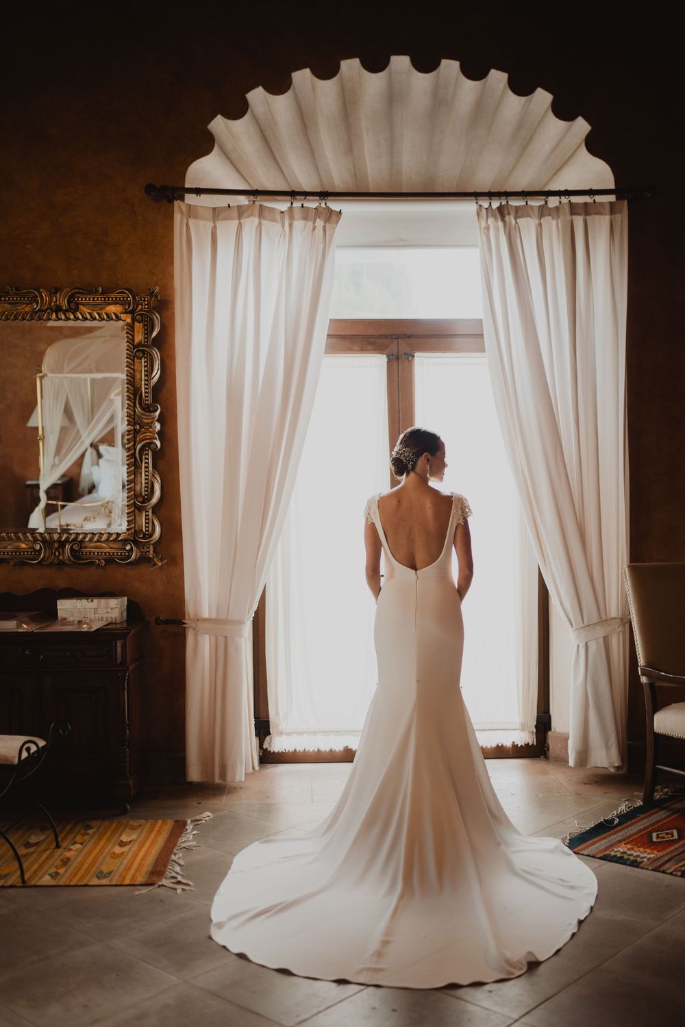 Elegant wedding dress silhouette against bright window with ornate curtain frame.
