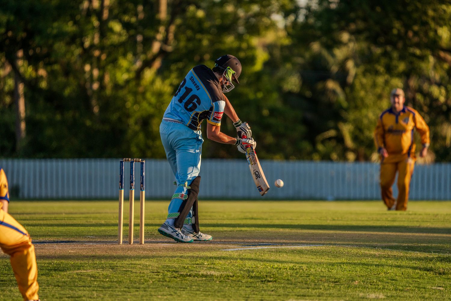 Cricket player in light blue uniform takes a powerful swing at the ball during a match at sunset.