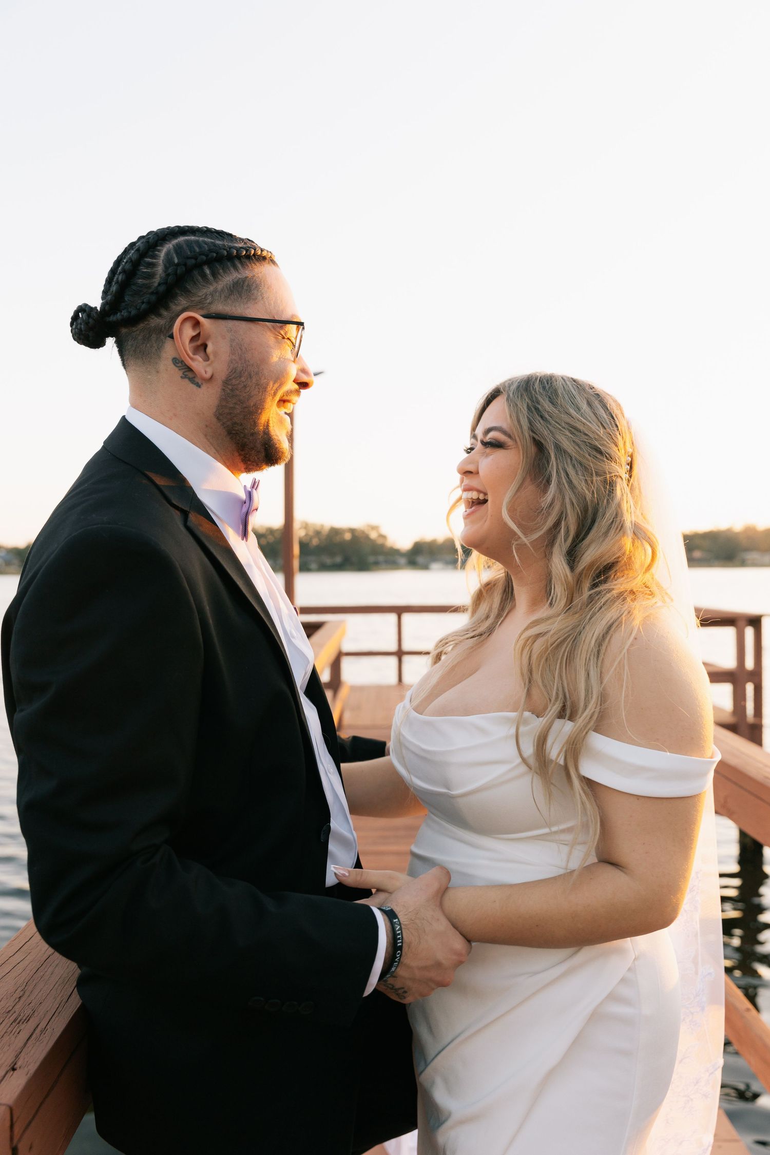 A laughing bride in an off-shoulder white gown and a groom with braided hair and glasses stand together on a waterfront dock at sunset.