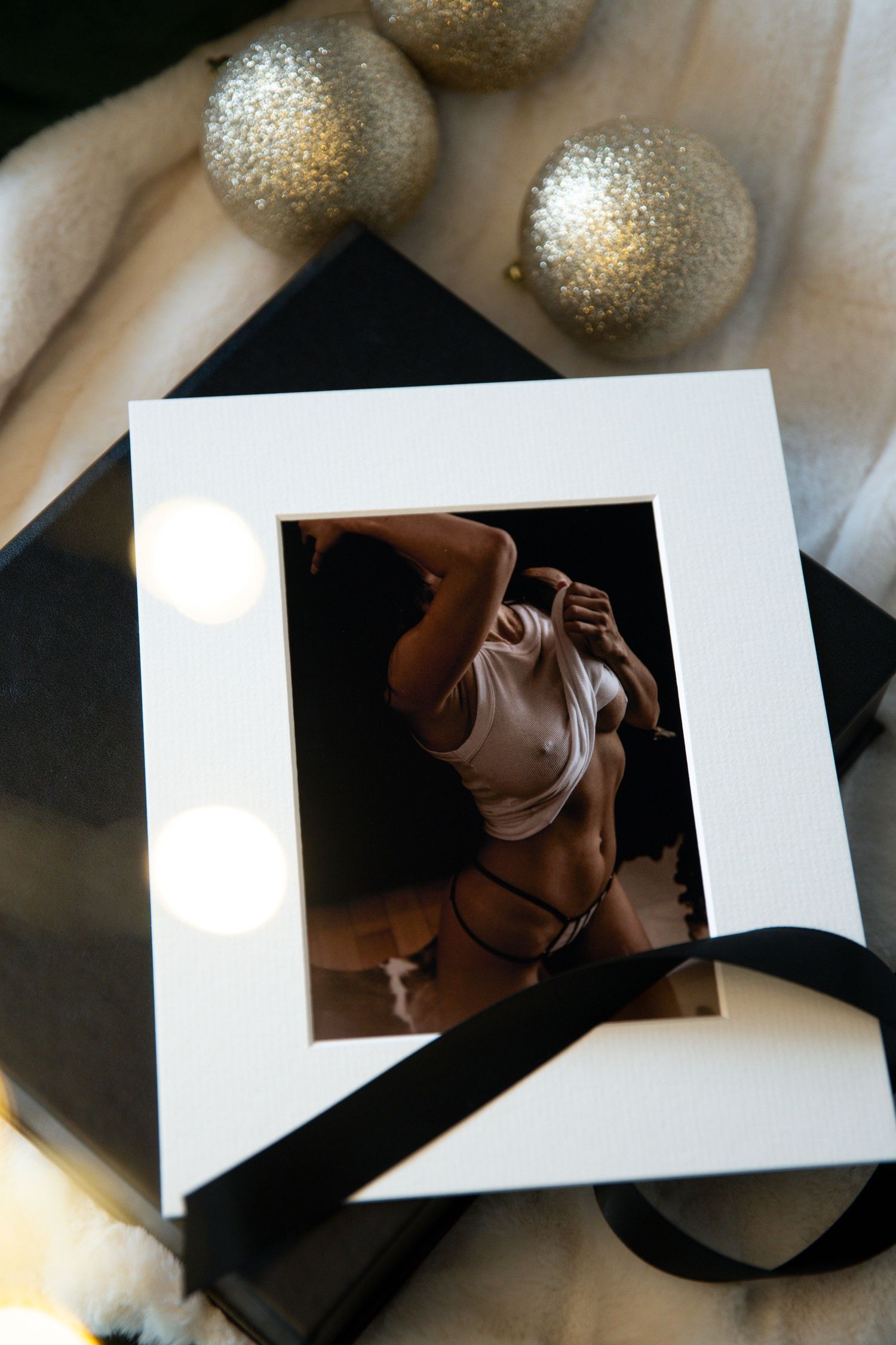 Artistic boudoir photo in white mat frame with silver glitter ornaments and black ribbon on light background.