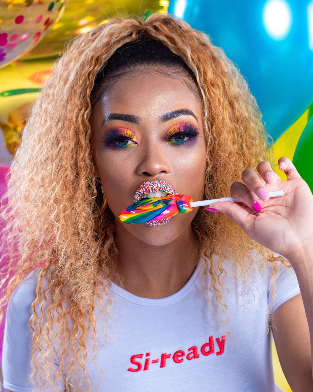 Vibrant beauty shot with blonde hair, rainbow makeup and a lollipop against colorful party backdrop.