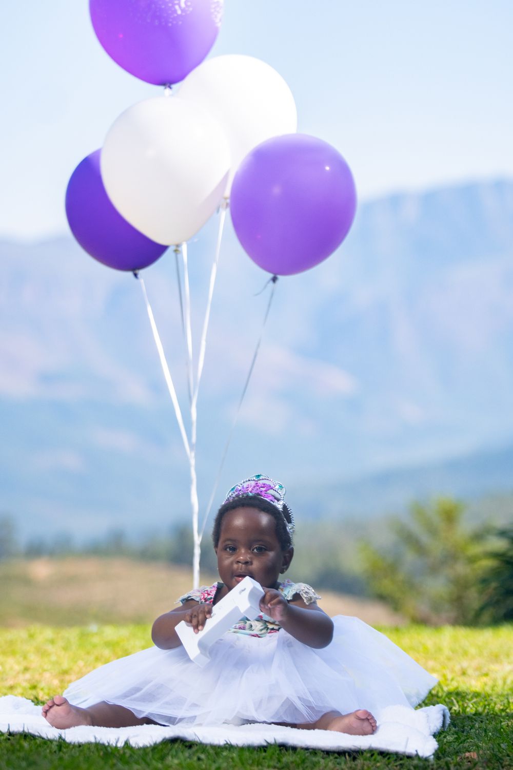 Child in white dress with tiara holds purple and white balloons against mountain backdrop.