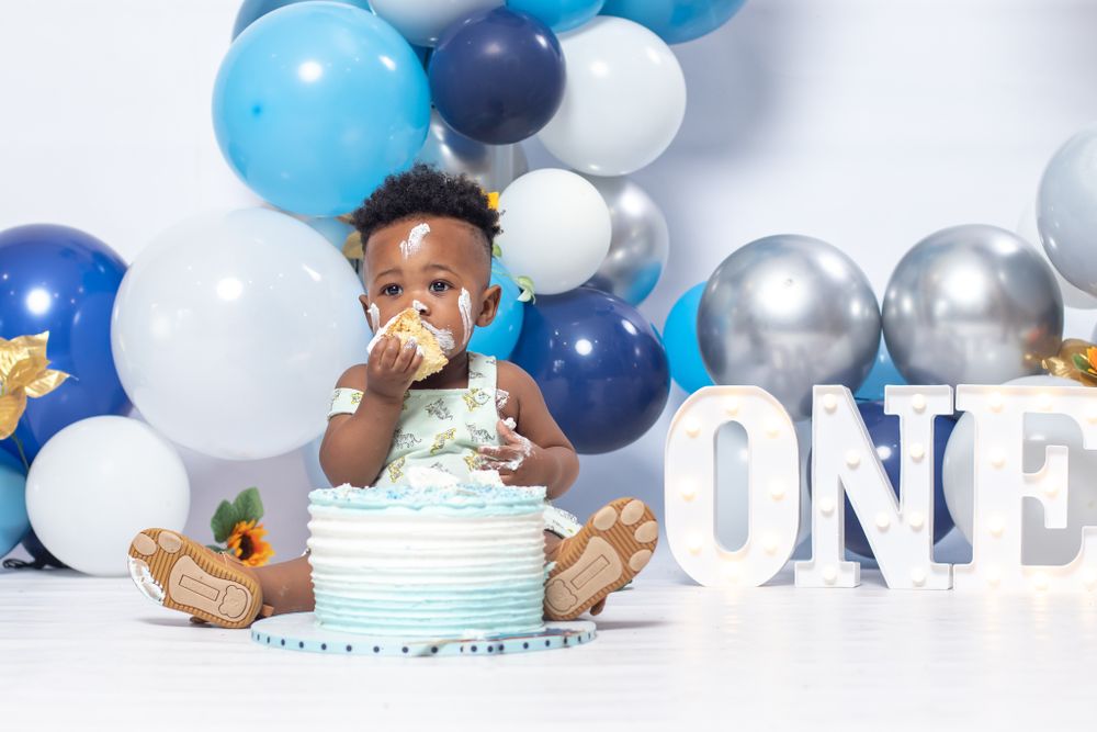 Baby enjoying birthday cake surrounded by blue and silver balloons.