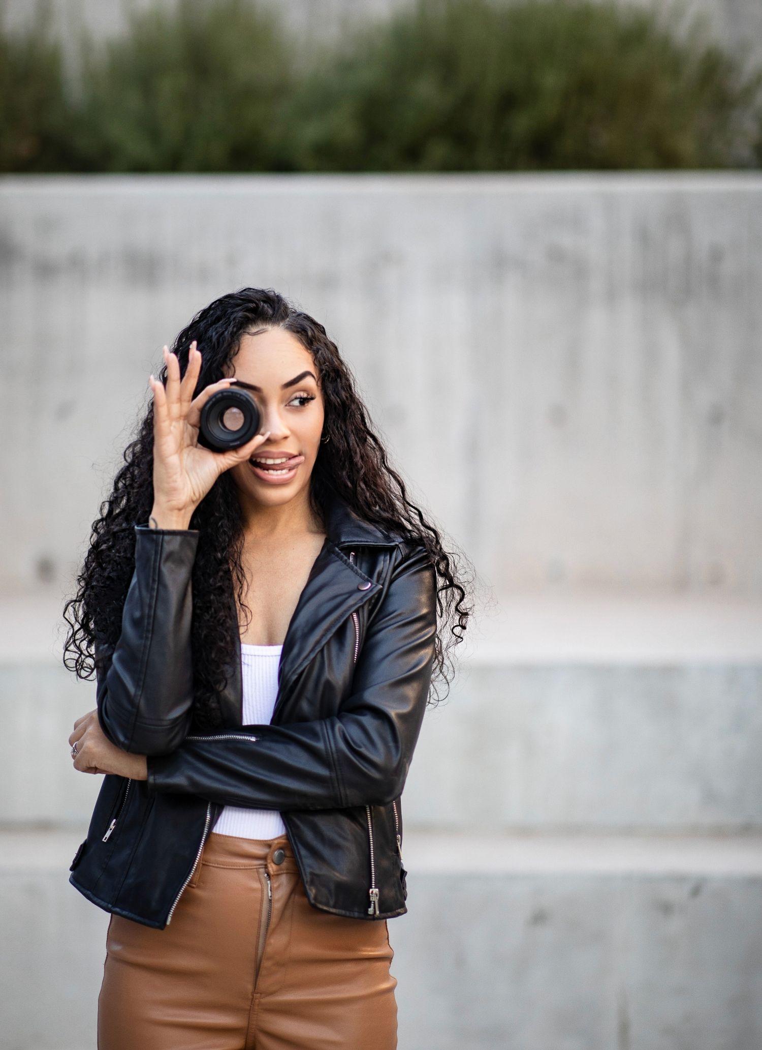 Creative photographer holds camera lens to eye while wearing leather jacket and brown pants.