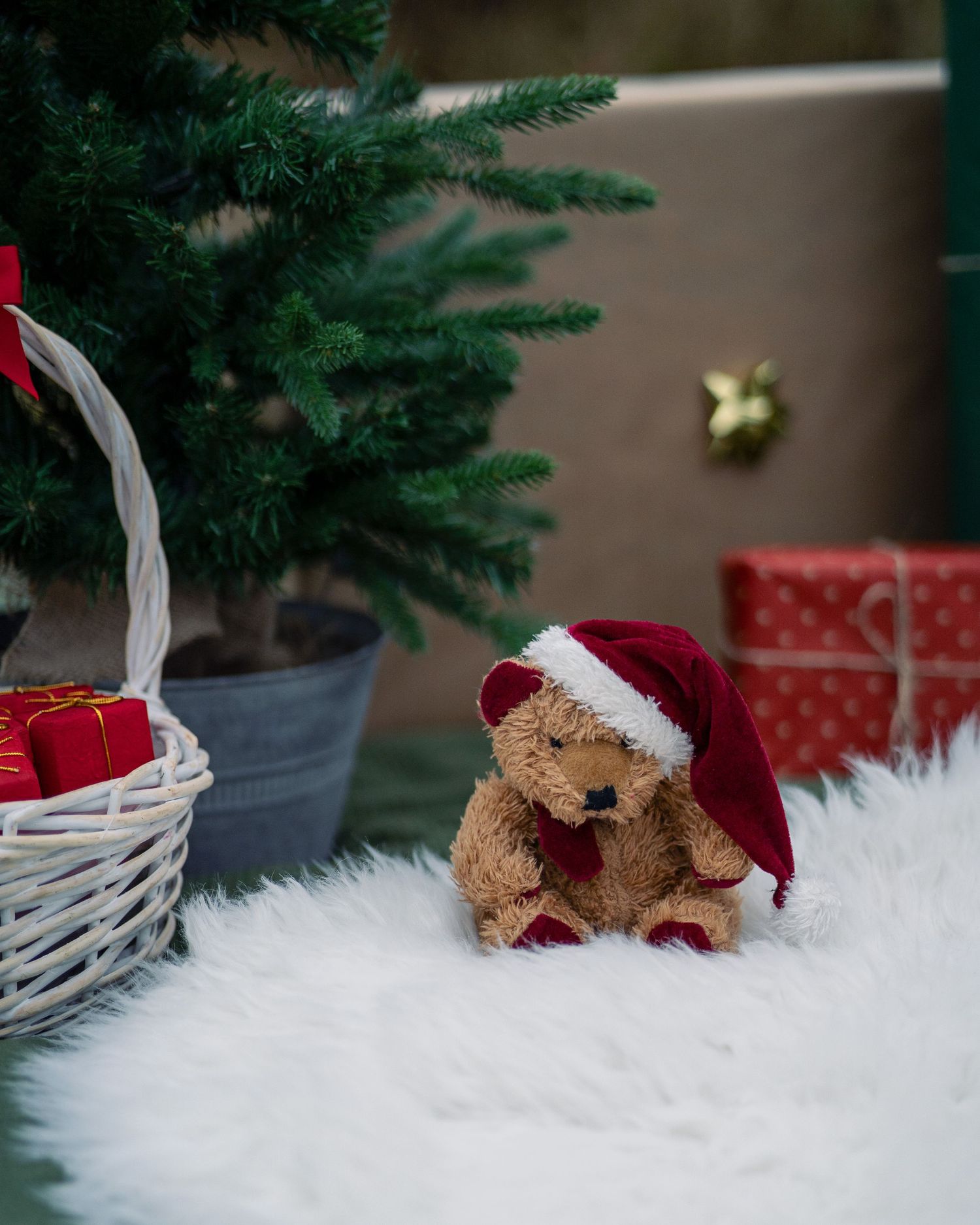 Festive display with plush teddy bear in Santa hat next to Christmas tree and gift basket.