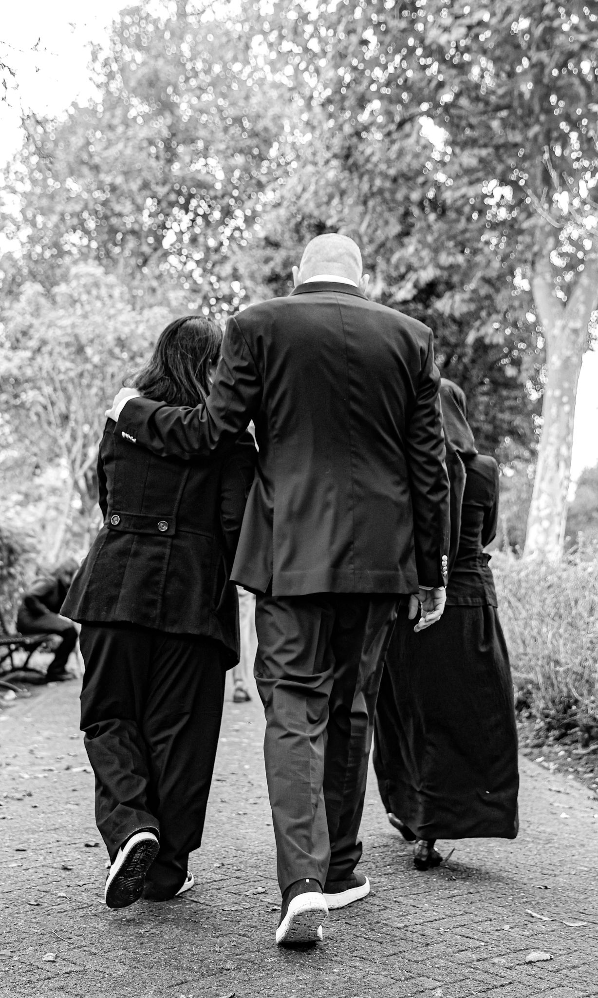 Two people in dark coats walking together on a path surrounded by trees in black and white.