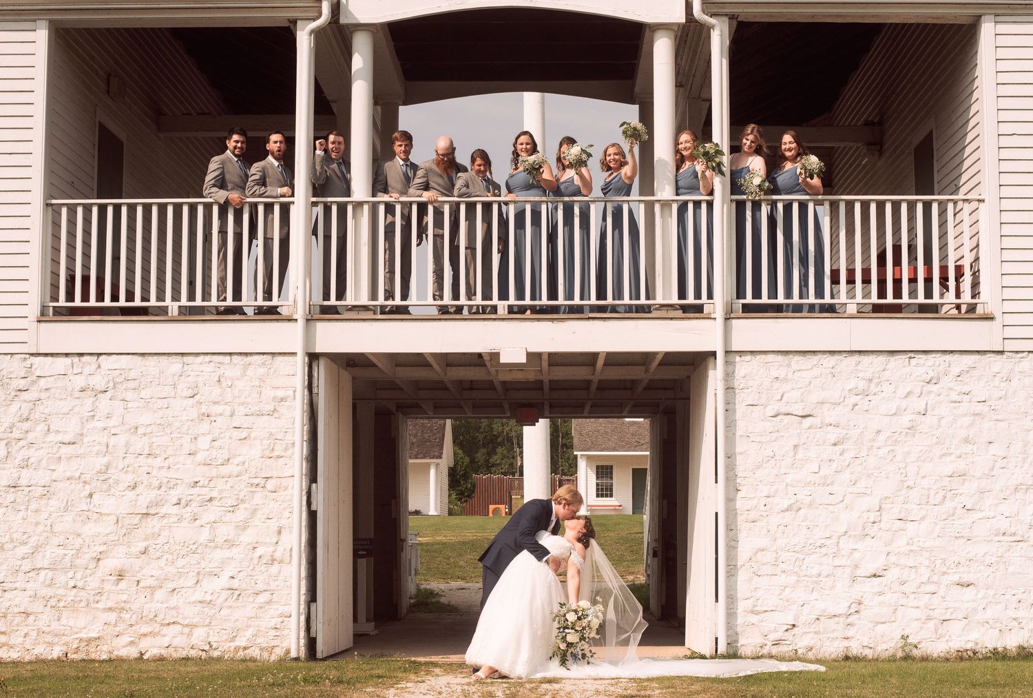 Bride and groom kissing under a balcony filled with wedding party guests at a rustic white building venue.