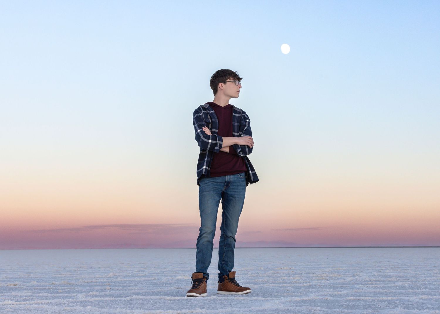 Person wearing plaid shirt and jeans stands for Bonneville salt flats photography at sunset as moon rises in pastel sky.