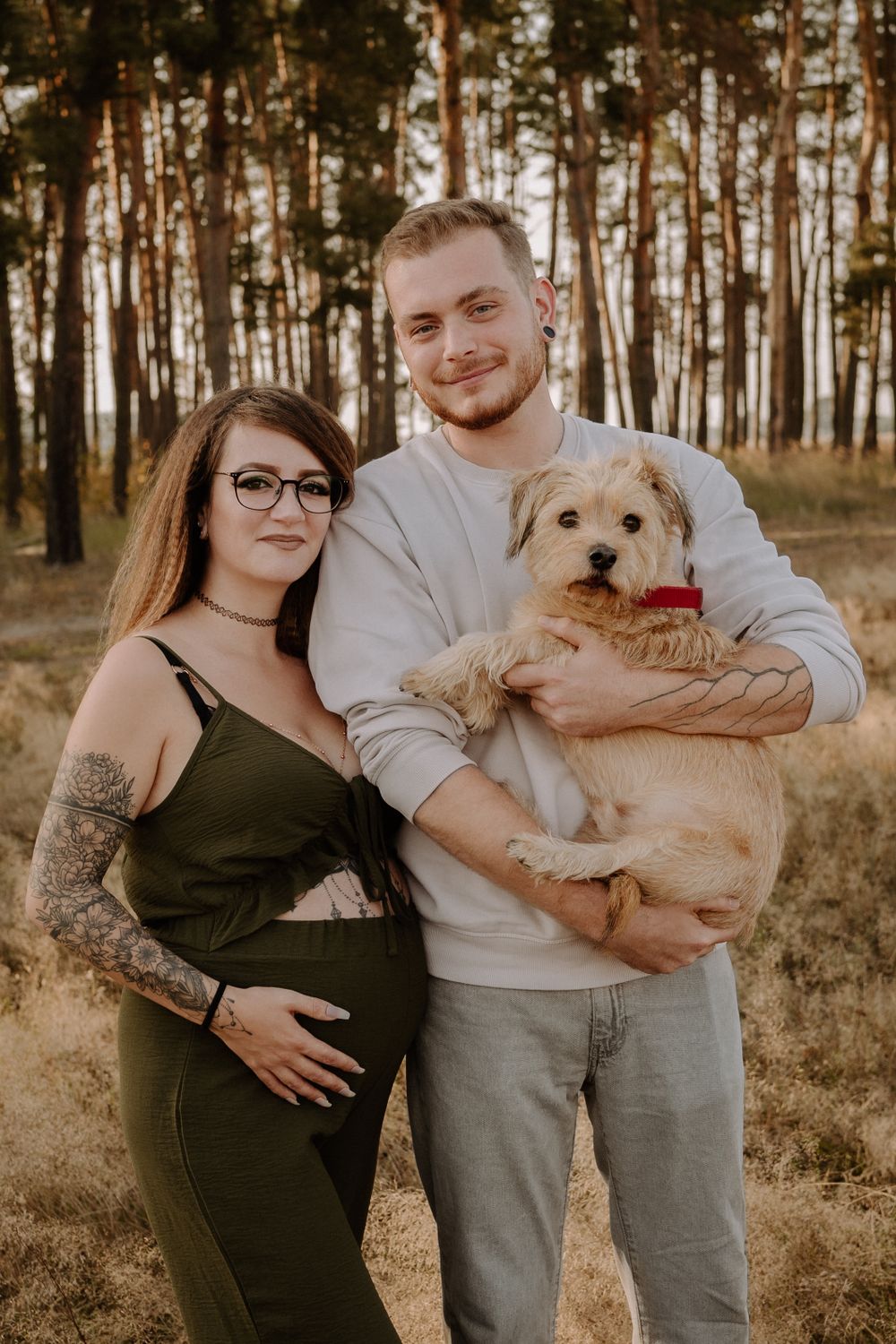 A couple stands in a pine forest during golden hour with their small tan dog wearing a red collar.