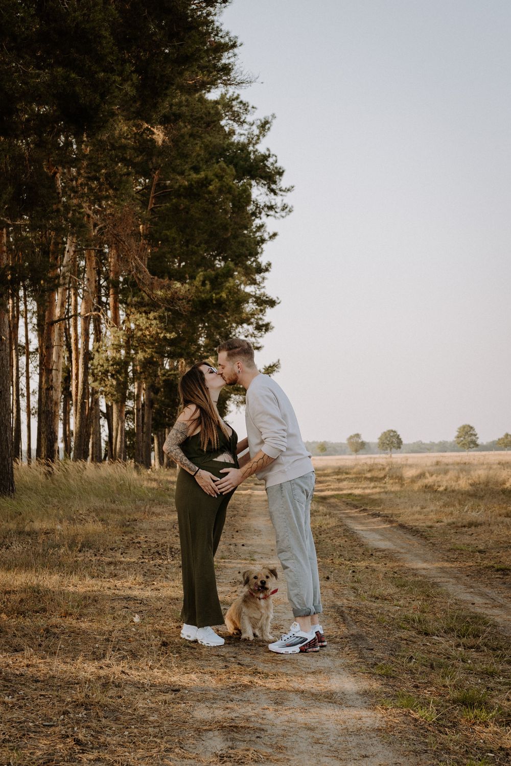 A couple stands on a dirt path through a pine forest with their small dog.