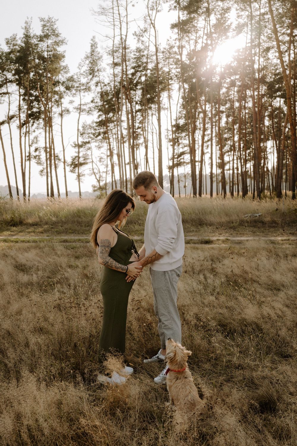 Couple stands together in a sunlit forest field with their dog at their feet.