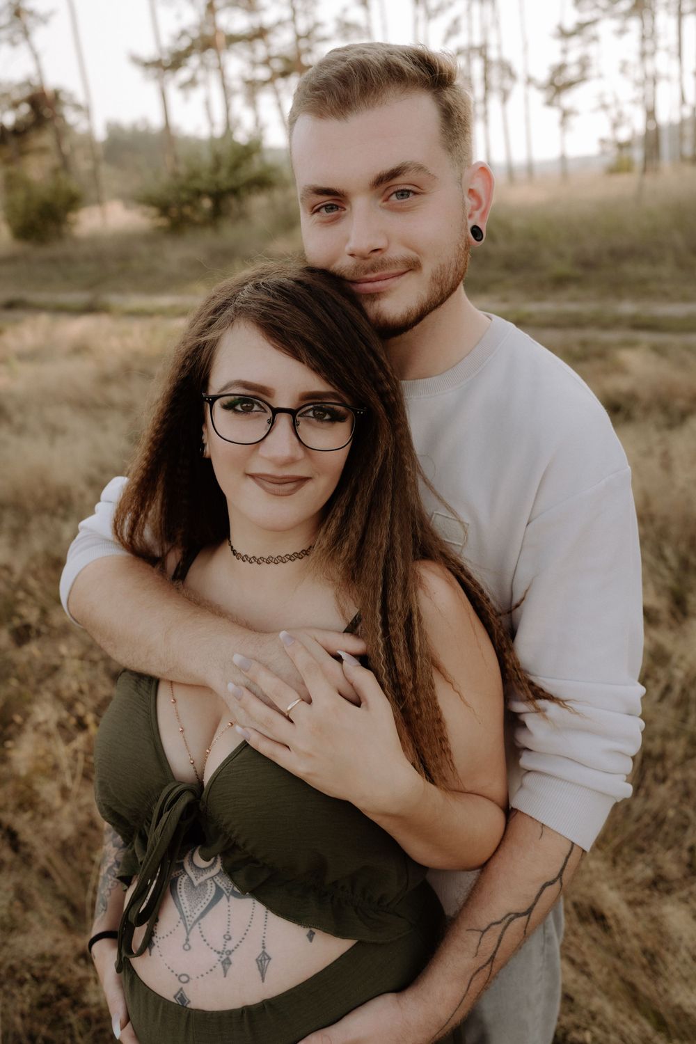 A couple embraces in an outdoor maternity photo shoot with pine trees in background.