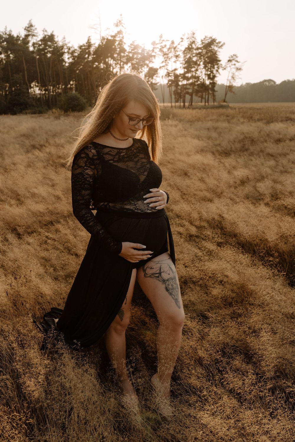 Expectant mother in black lace dress poses in golden field at sunset.