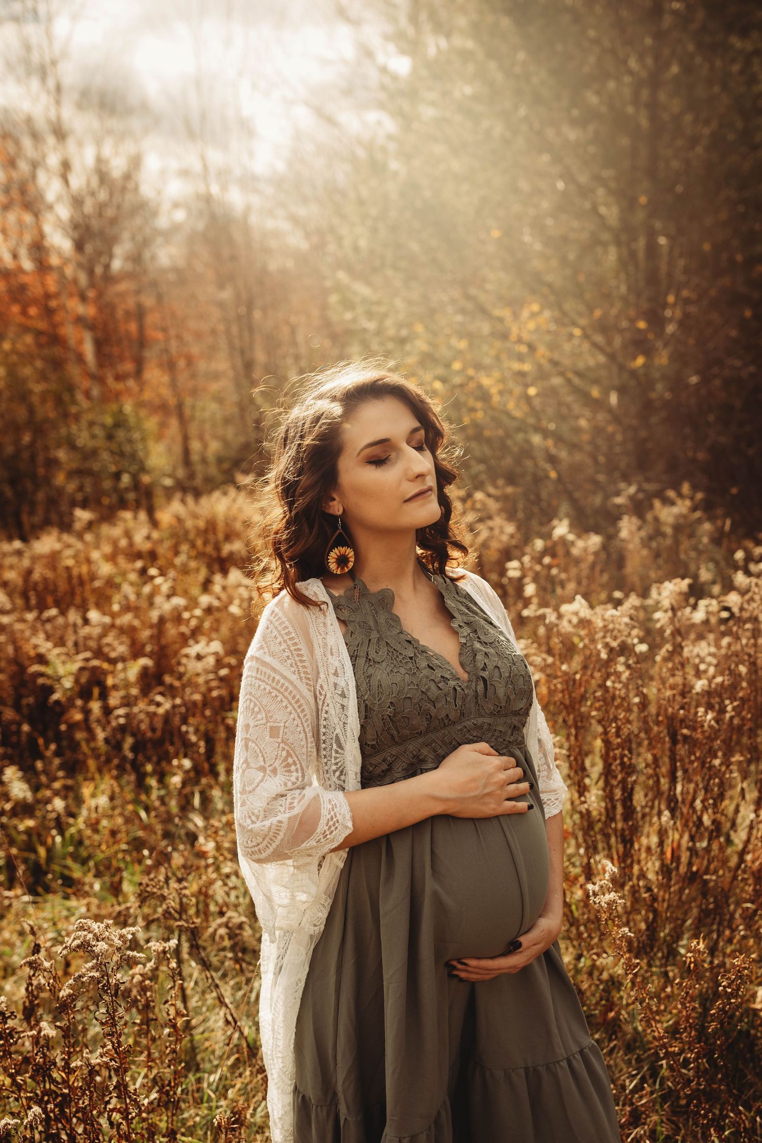 A maternity portrait in a grey dress and white cardigan set against autumn foliage.