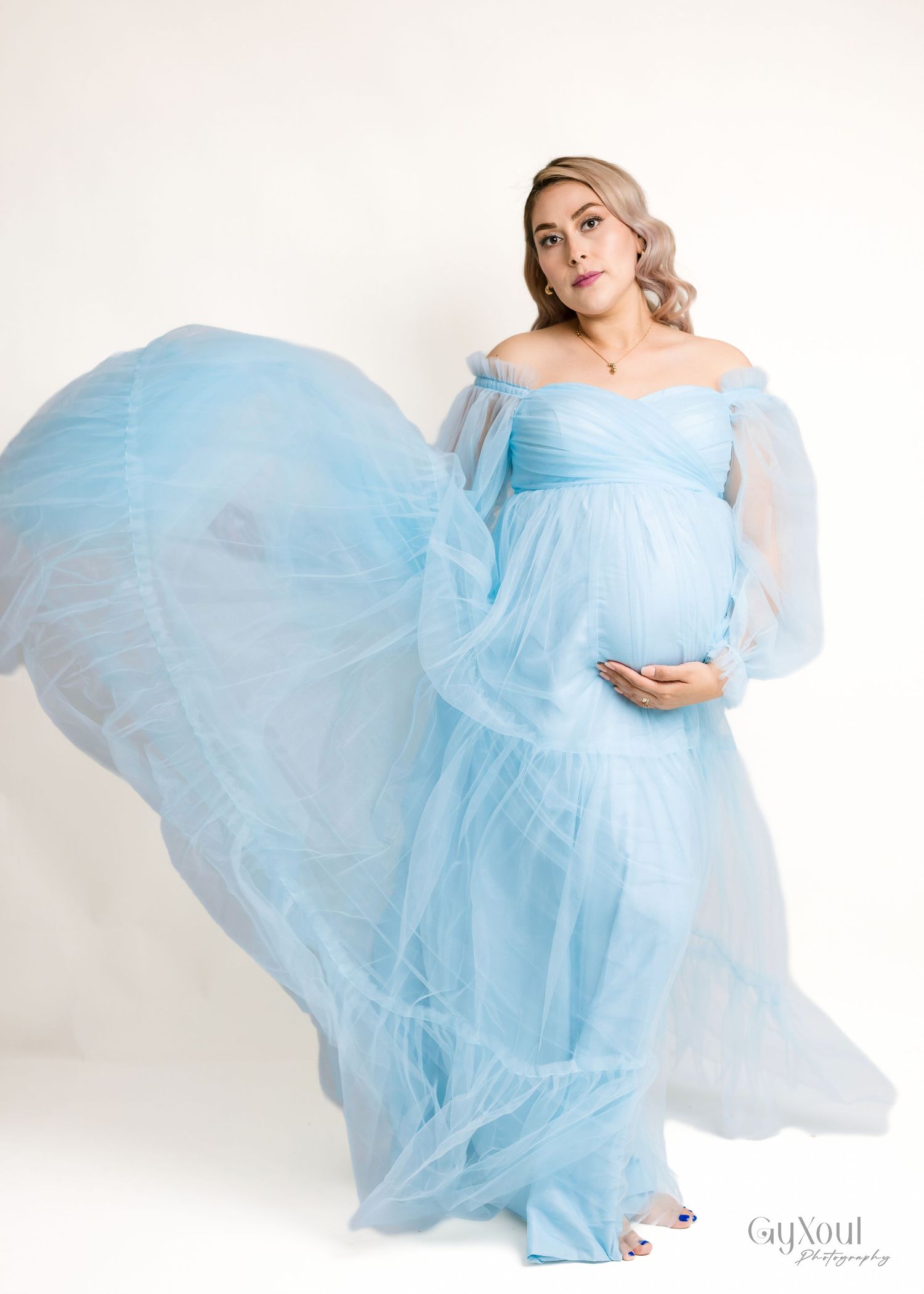 A woman in a flowing powder blue maternity gown twirls gracefully against a white studio backdrop.