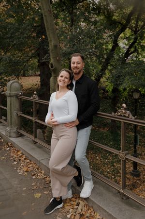 A couple poses together on a bridge railing surrounded by autumn trees in a park setting.