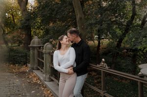 A romantic outdoor photoshoot shows a couple embracing on a wooden bridge surrounded by autumn foliage.