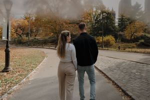 A couple walks together on a curved path surrounded by autumn trees and fallen leaves on a misty day.