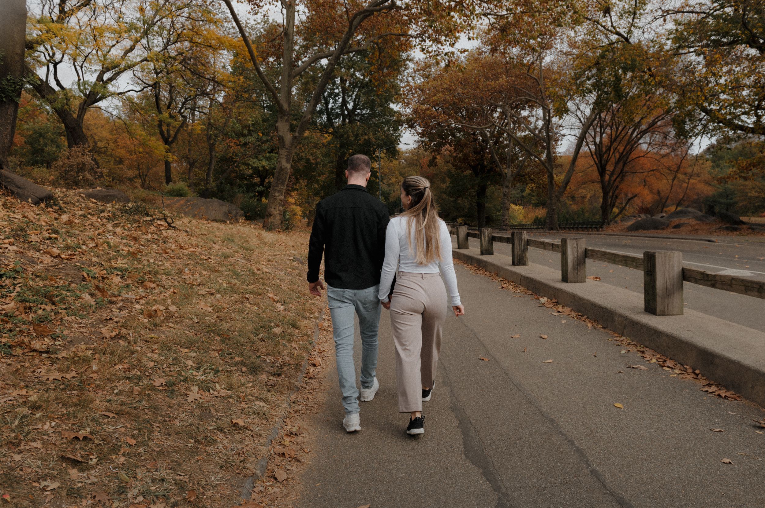 A couple walks along an autumn path covered in fallen leaves in a park setting.