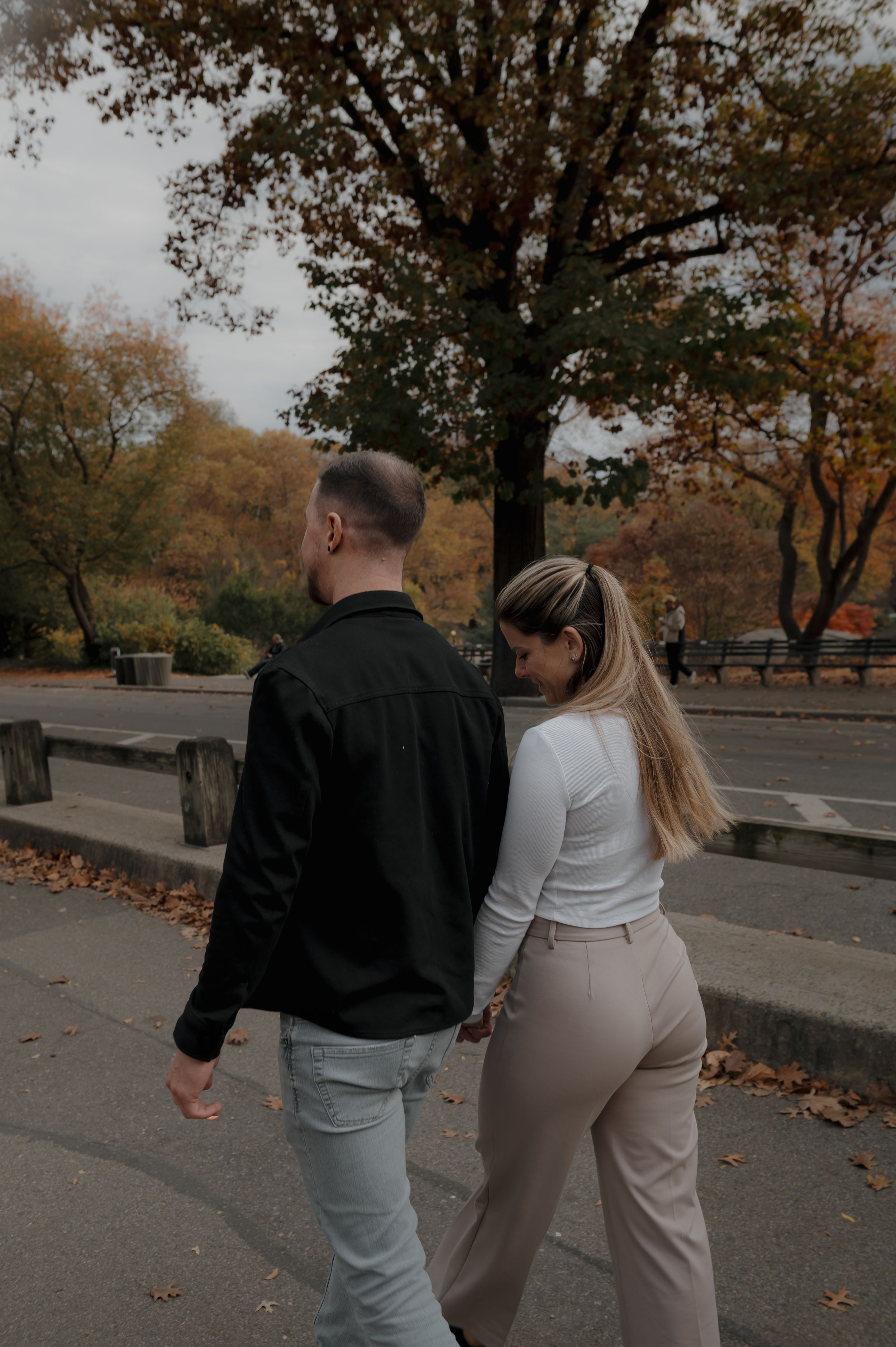A couple walks together on an autumn path with fallen leaves and trees in the background.