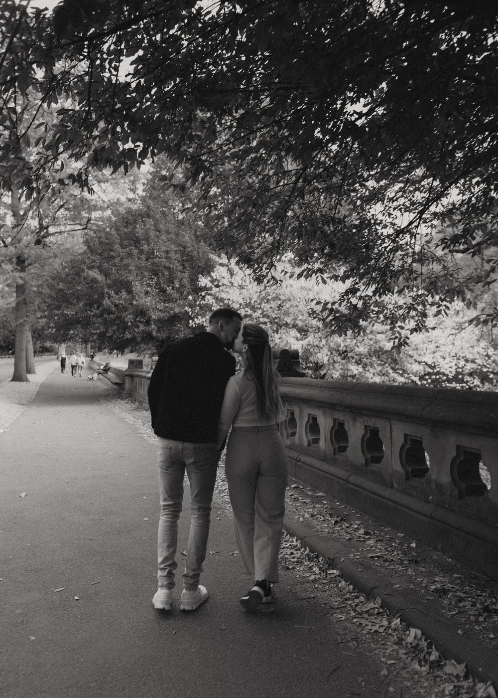 Two people stroll down a tree-lined path with a stone railing during fall.