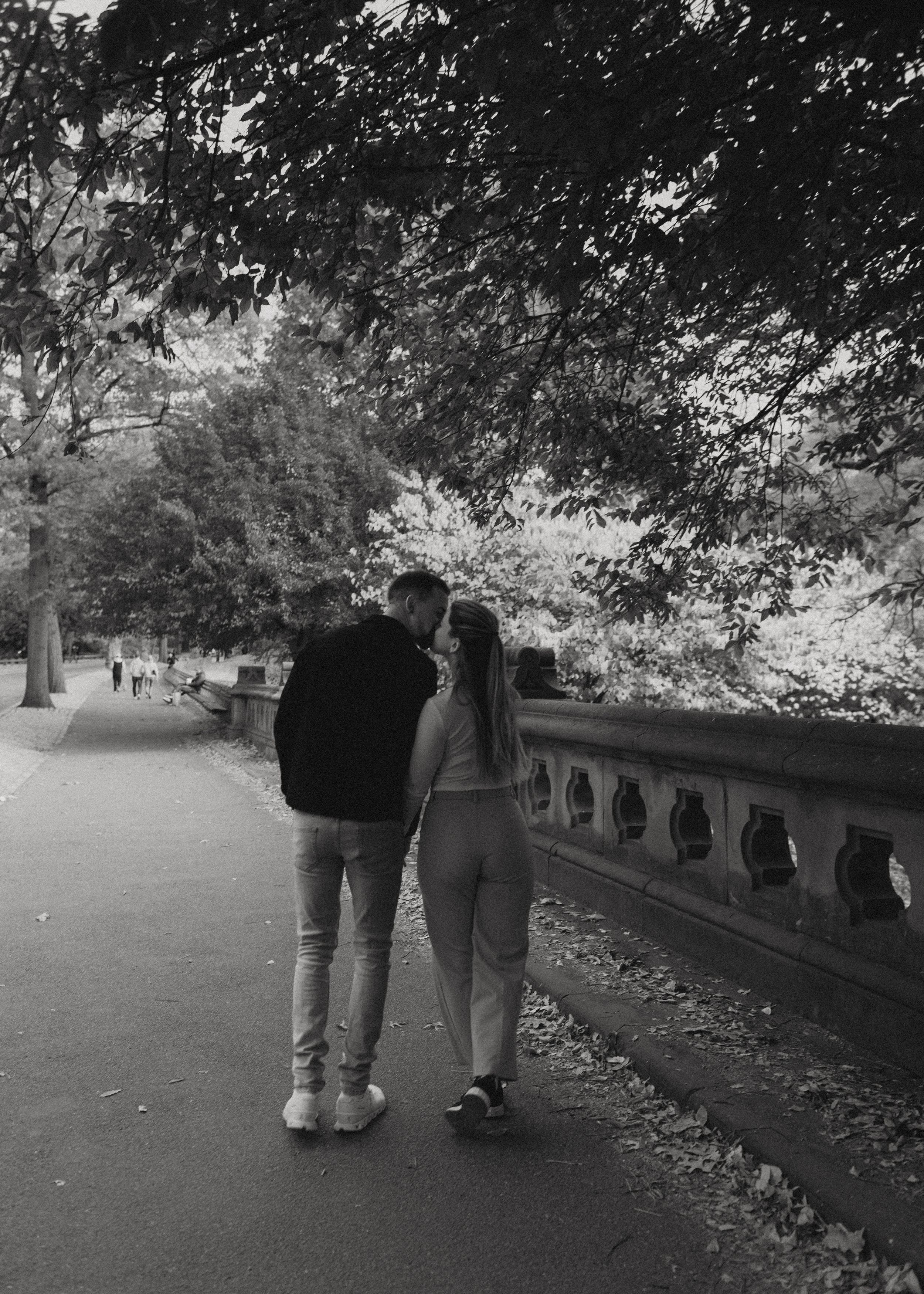 Two people stroll down a tree-lined path with a stone railing during fall.