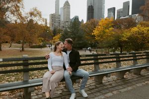 A couple shares an intimate moment on a park bench with NYC skyscrapers and fall foliage in the background.