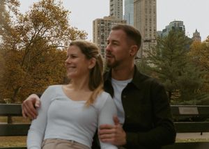 A couple embracing and smiling against an urban backdrop of tall buildings and fall foliage in a city park.