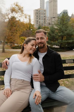 A couple shares an intimate moment while sitting on a park bench with autumn trees and city buildings in the background.