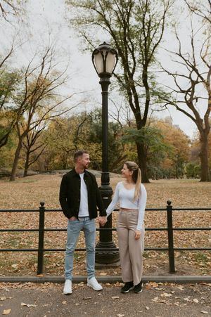 A couple sits on a park bench near a vintage lamppost surrounded by fallen autumn leaves.