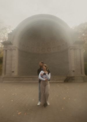 Two people embrace in front of a misty stone archway during fall.