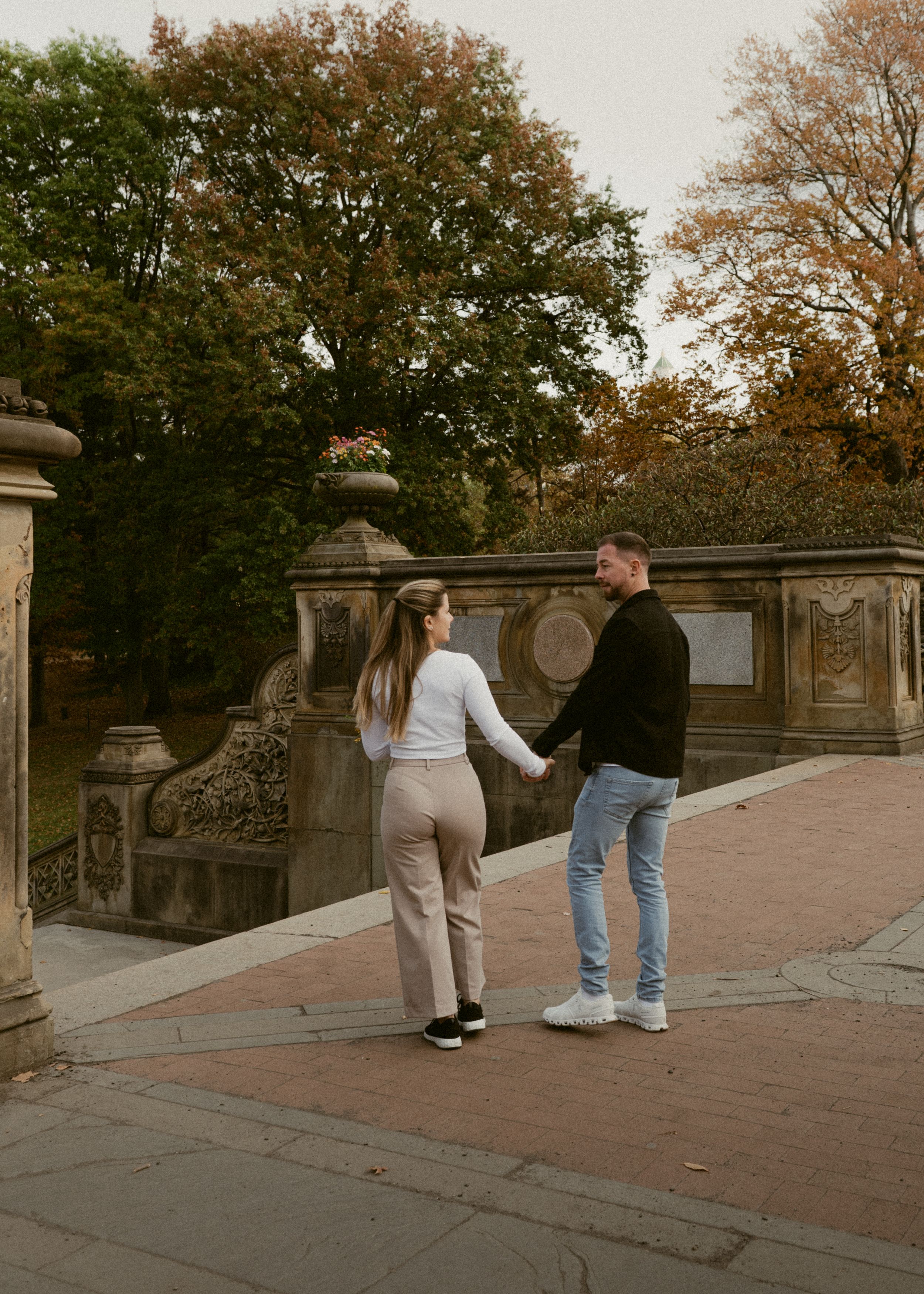 A couple walking hand in hand on a stone pathway surrounded by autumn trees and ornate railings.