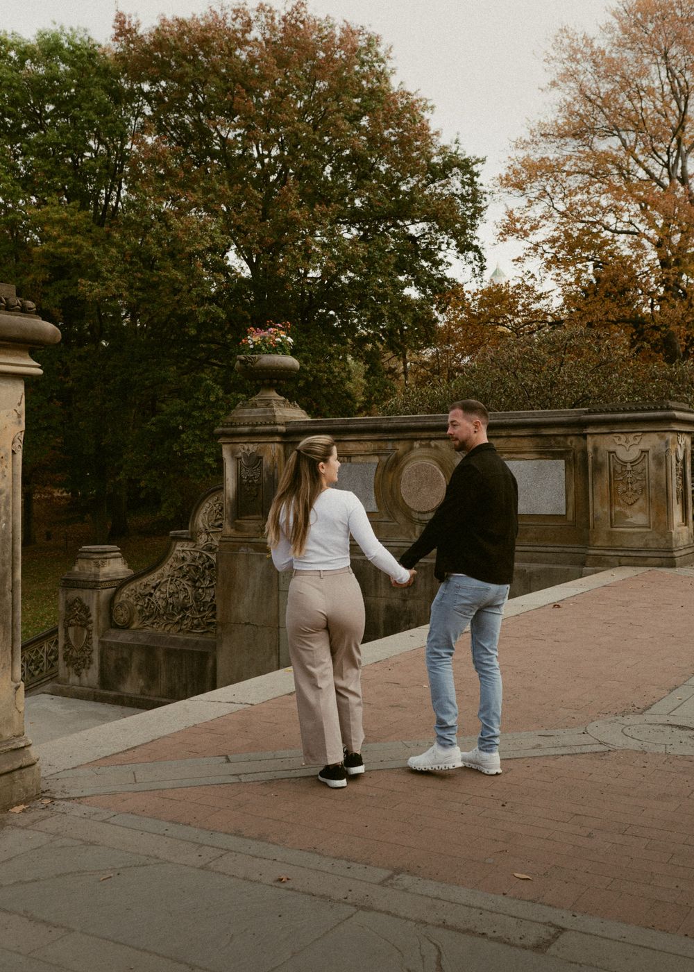 A couple walking hand in hand on a stone pathway surrounded by autumn trees and ornate railings.