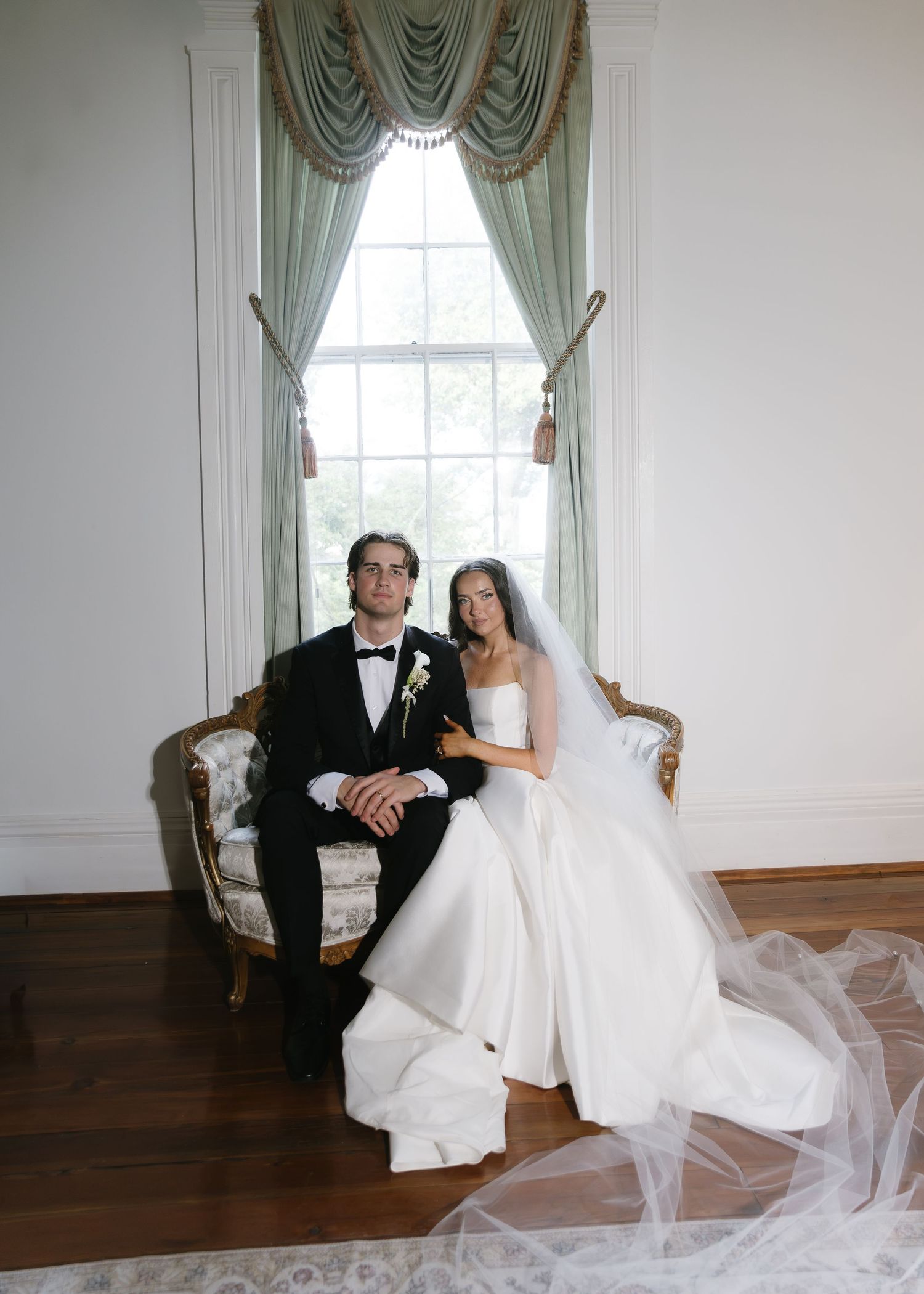 Elegant bride and groom sitting on ornate gold settee in white ballroom, bride in strapless gown with veil, groom in black tuxedo.