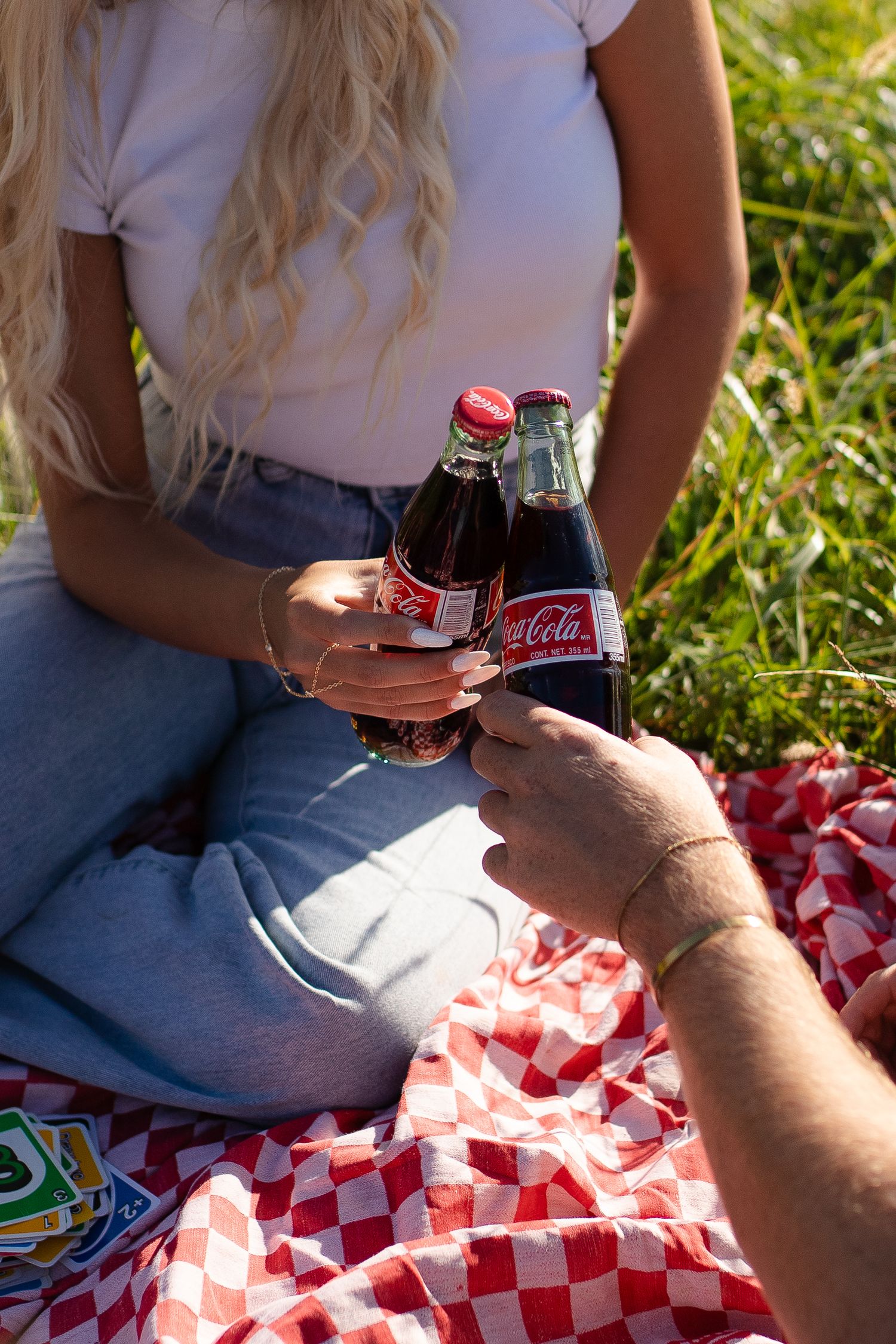 Hands holding Coca-Cola bottles on a red and white checkered picnic blanket. Couples photo session