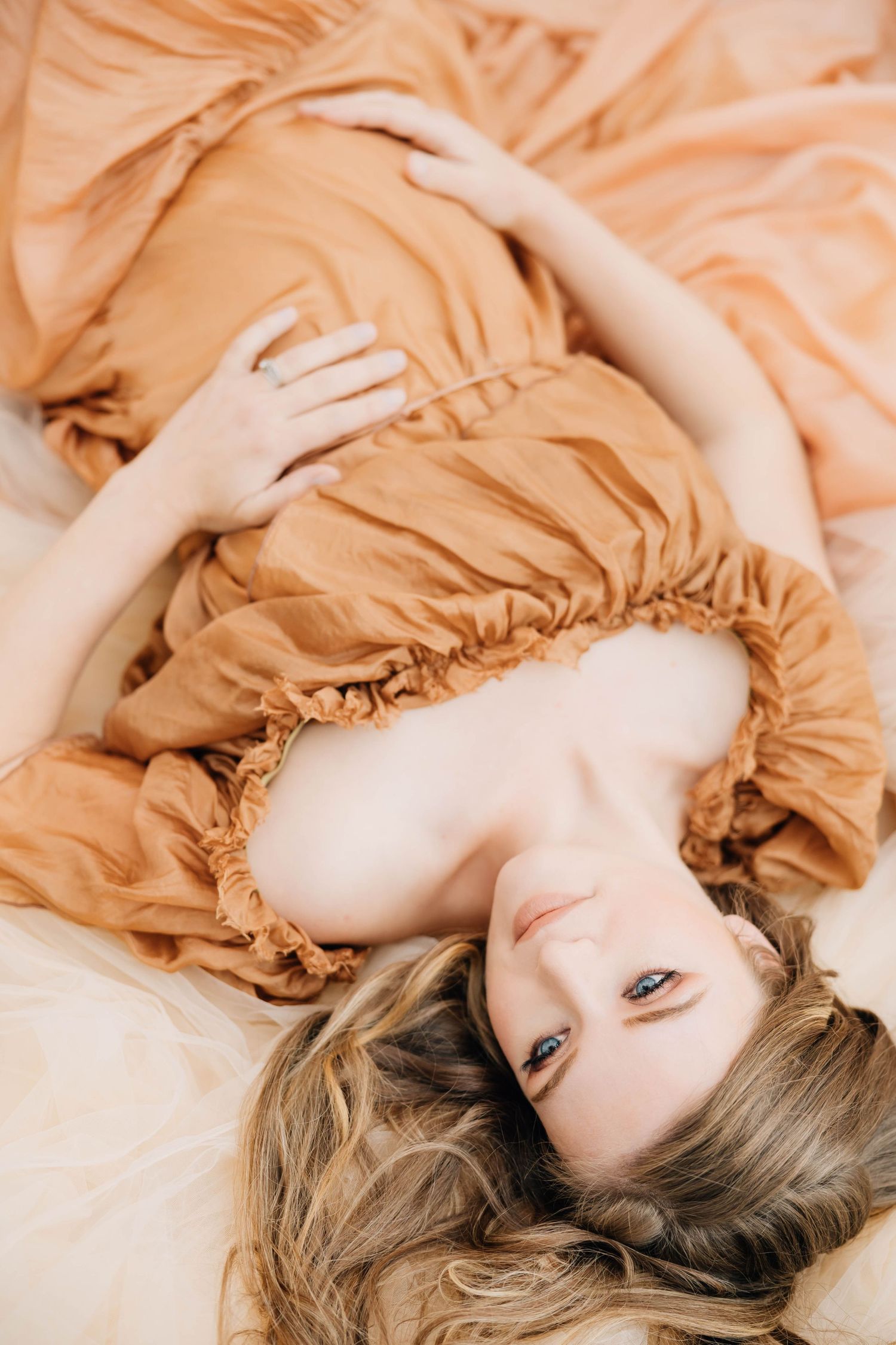 Pregnant woman in rust orange dress lying down looking up at camera with flowing hair.