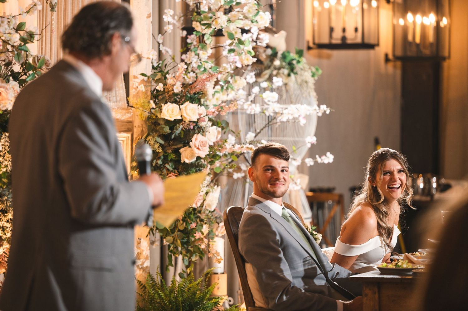 Couple laughs during toast seated at elegant farm table with flowers and warm rustic lights at The Barn at Villa Venezia Middletown, NY.