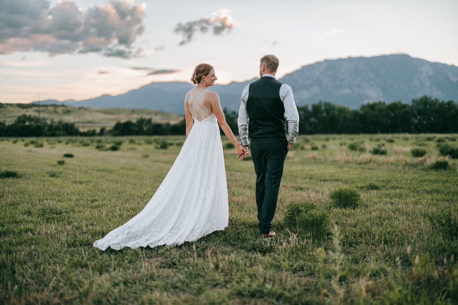 A bride and groom walking together across a scenic mountain meadow at sunset.
