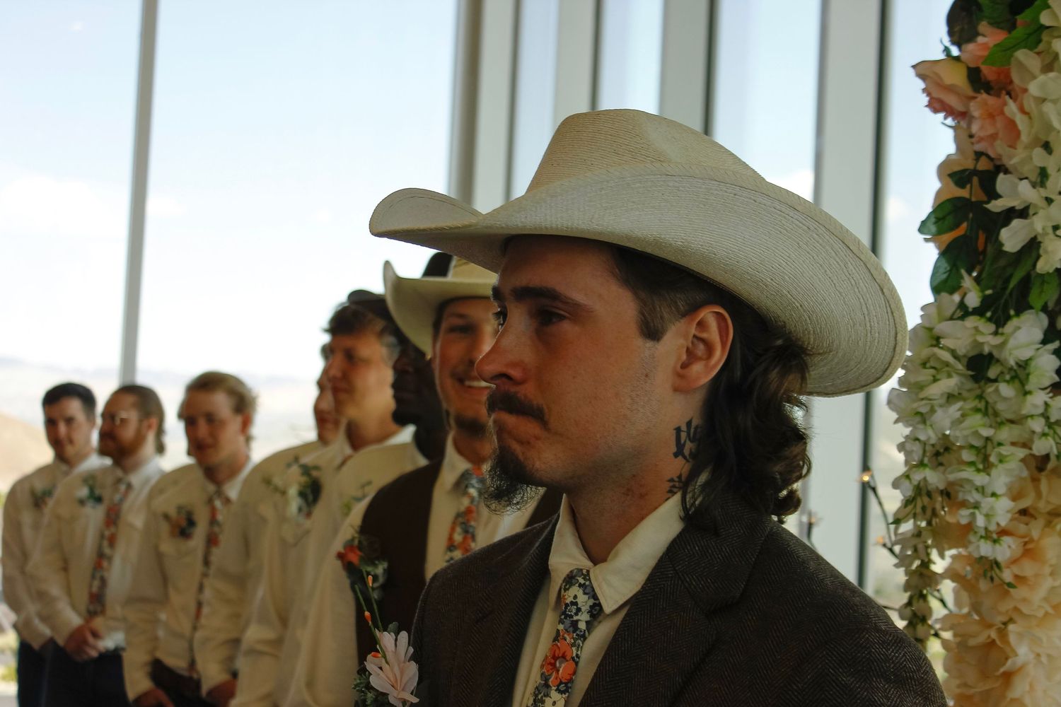 Groom in white cowboy hat and floral tie stands at wedding altar with groomsmen in tan suits behind him.