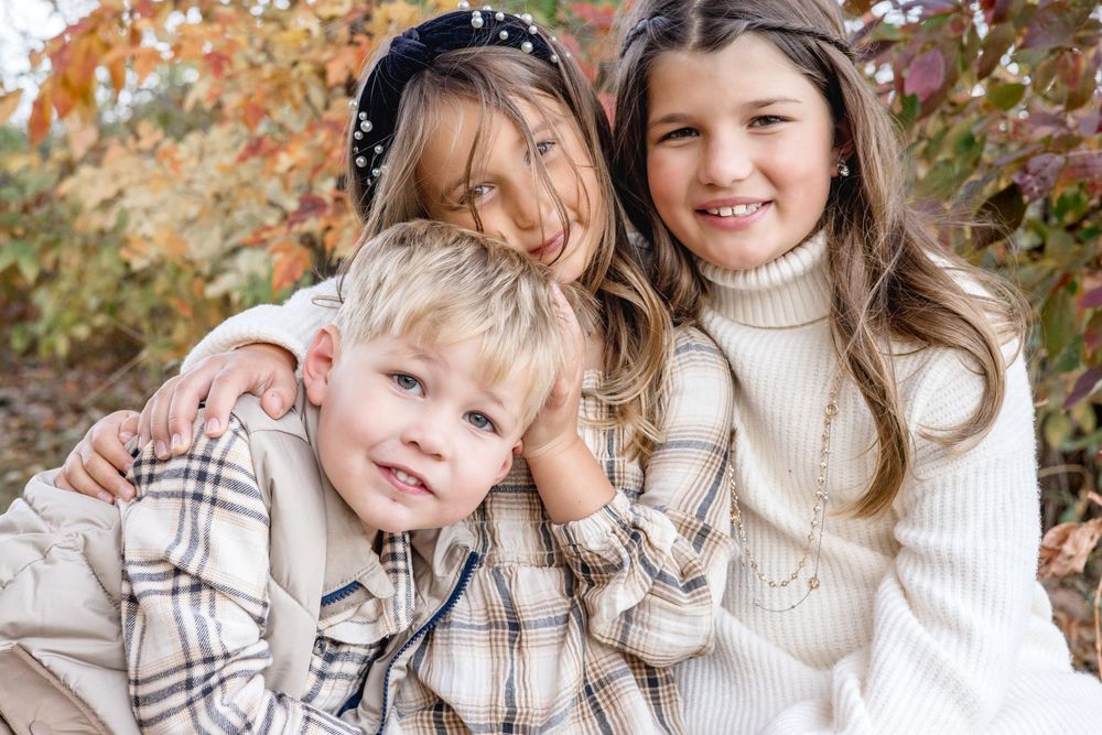 Three children in cream sweaters smiling together against autumn foliage.