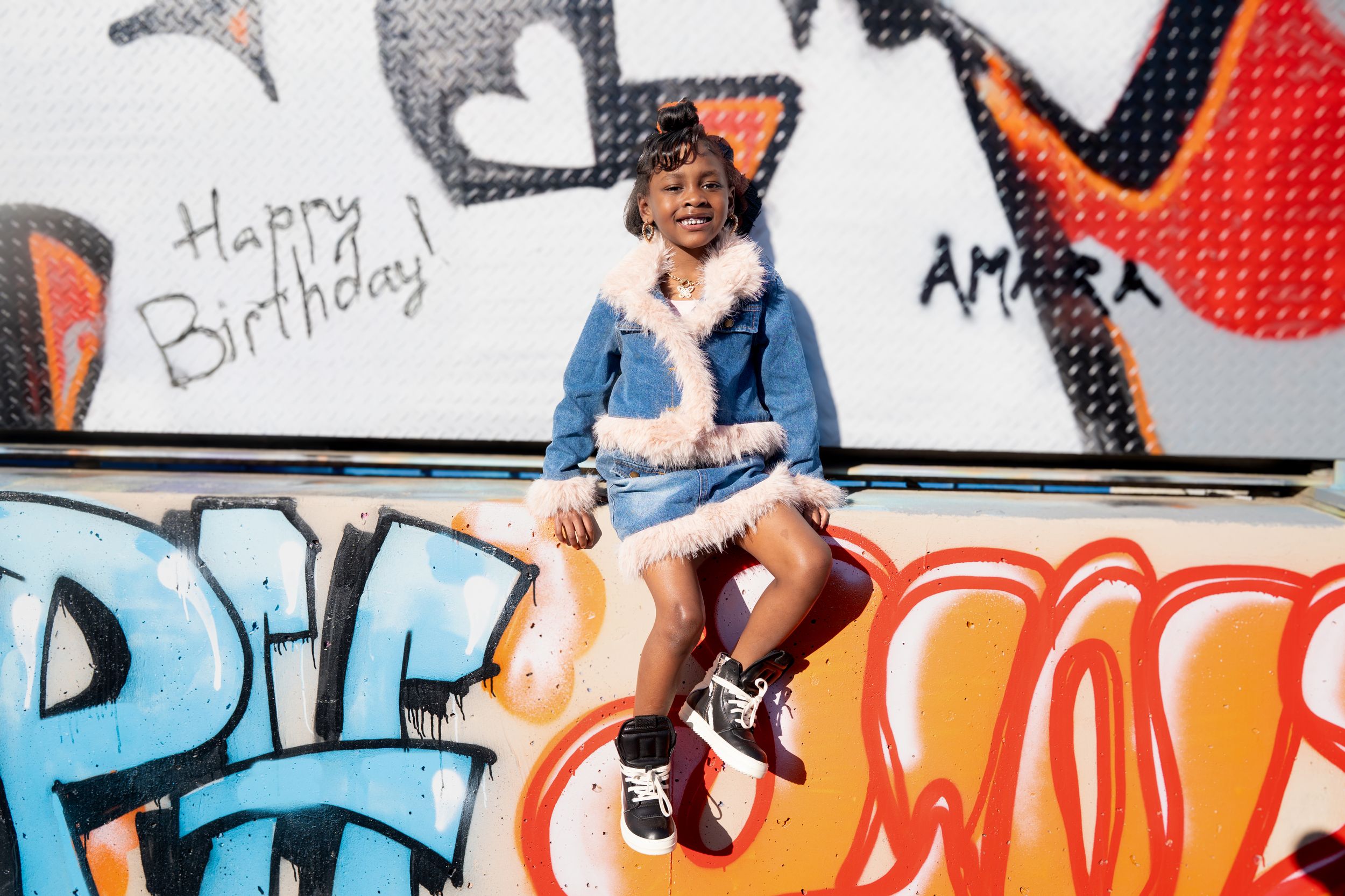 Someone sits casually against a colorful graffiti wall with 'Happy Birthday' written on it, wearing denim and white outfit.