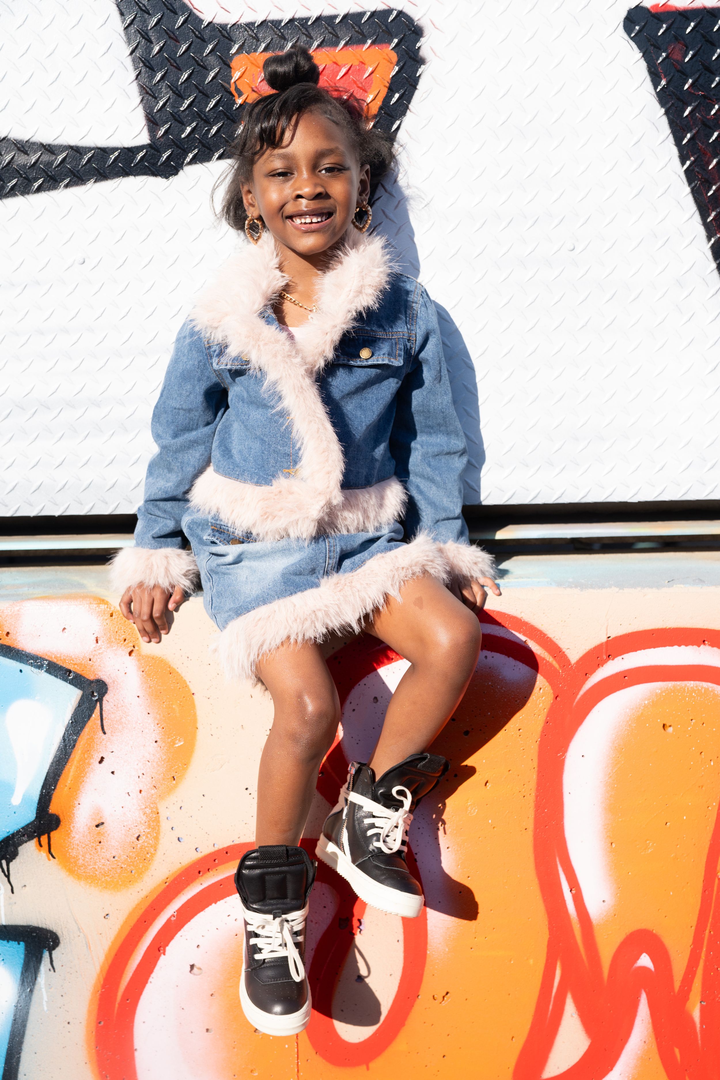 A child wearing a denim jacket with white fur trim and black sneakers sits playfully on a colorful graffiti wall.