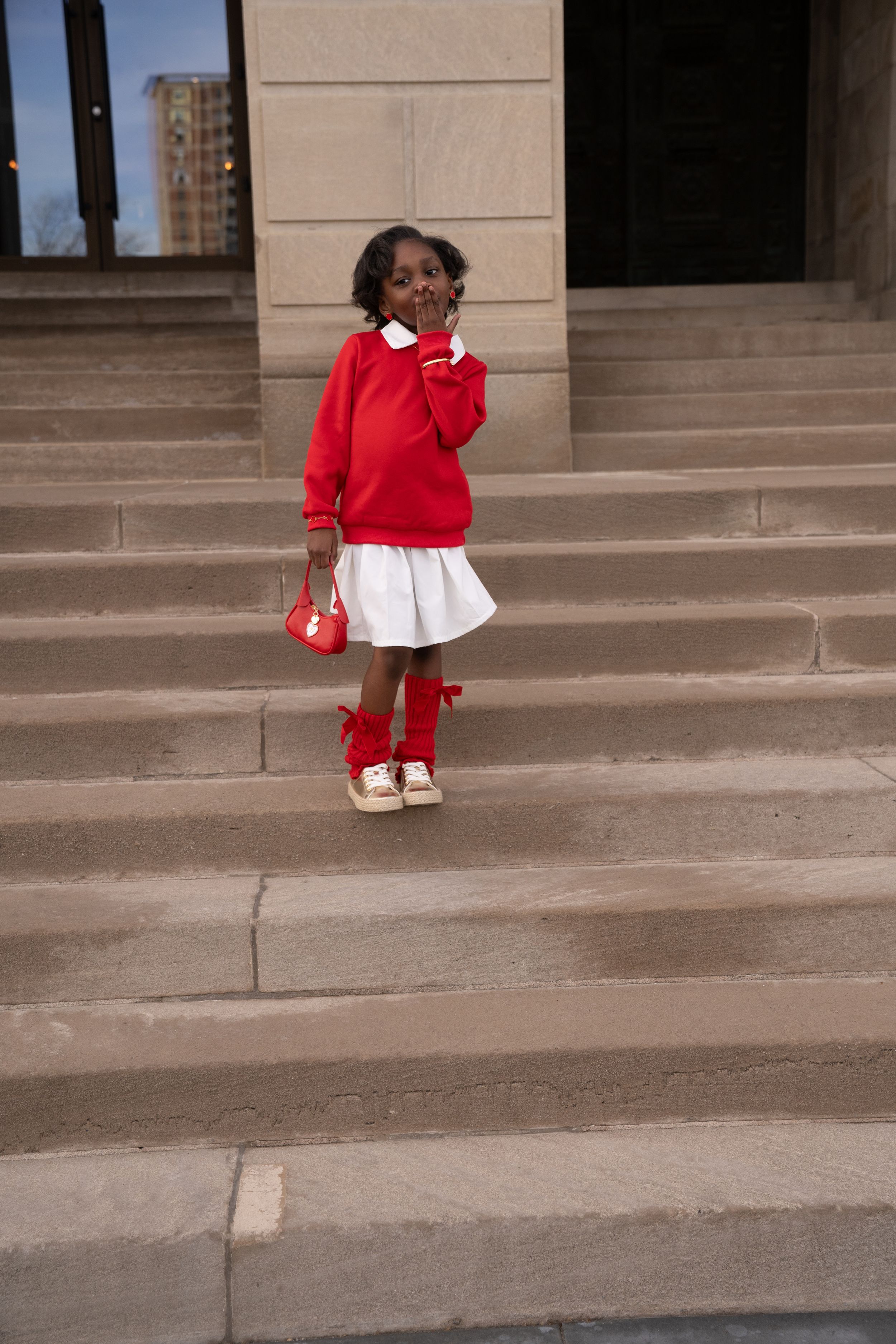 A stylish young child stands on grey stone steps wearing a bright red sweater, white skirt, and matching red shoes.