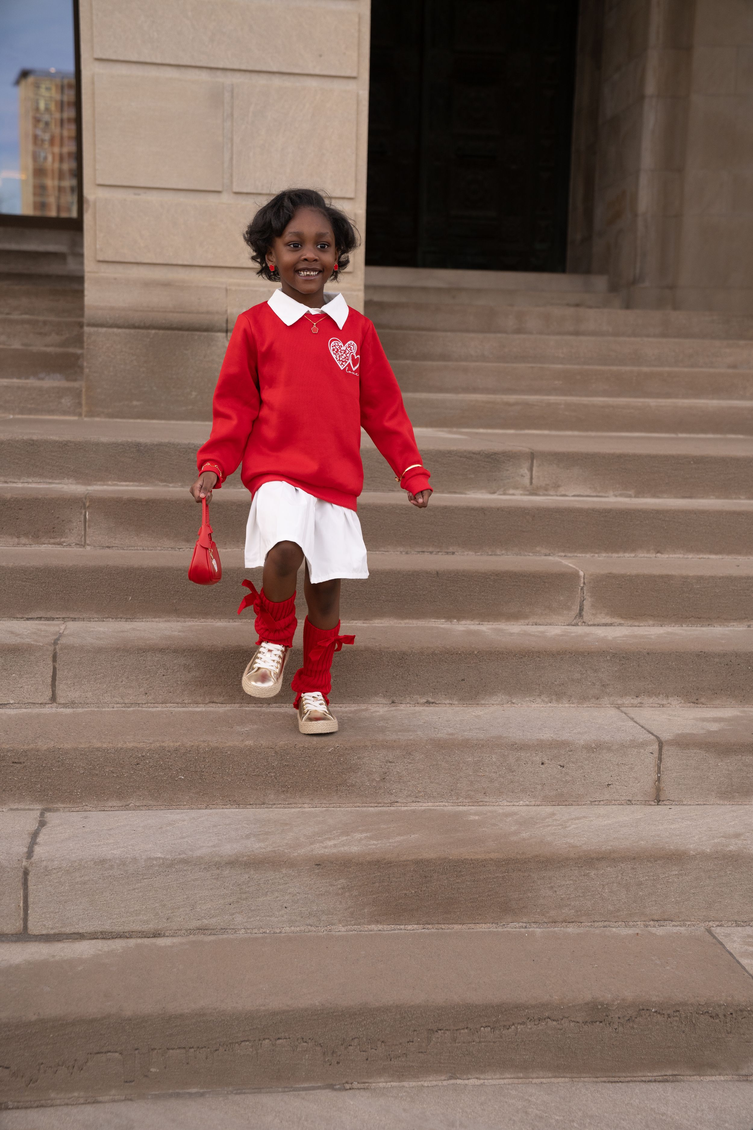 A child in a red sweater and white skirt walks down stone steps while wearing red knee socks and sneakers.