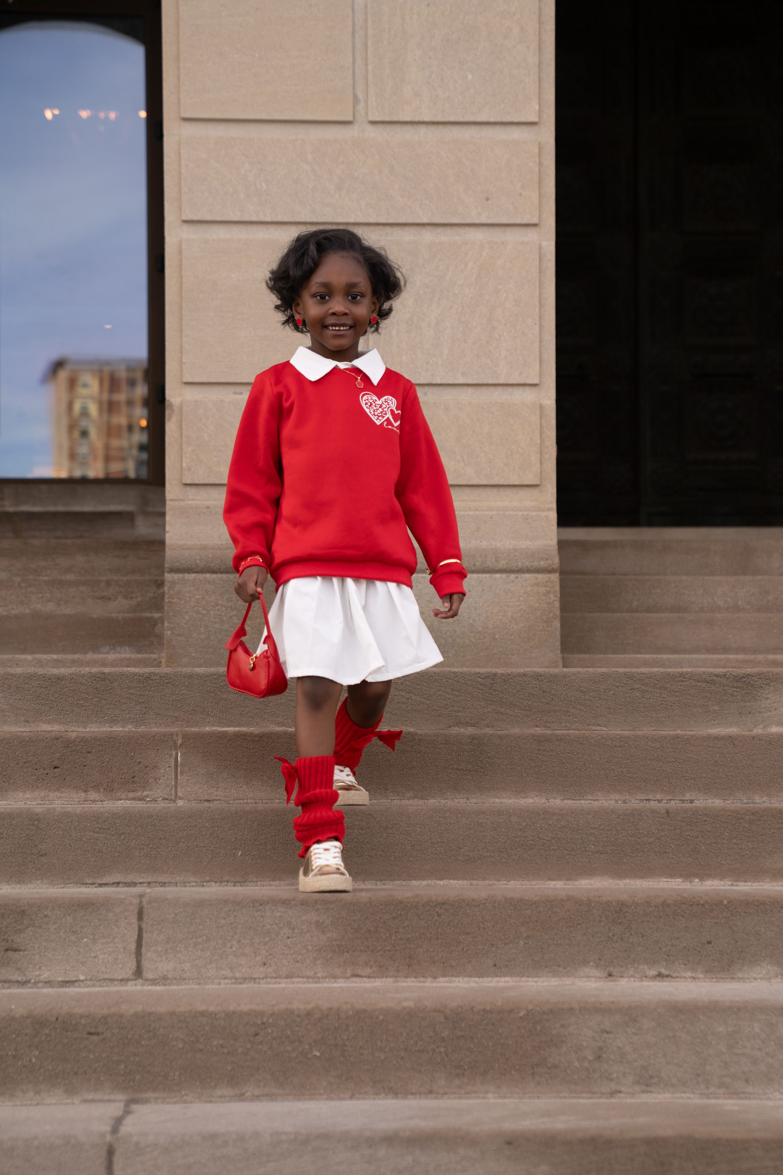 A student wearing a red sweater, white skirt, and red knee socks stands on stone steps with a matching red bag.