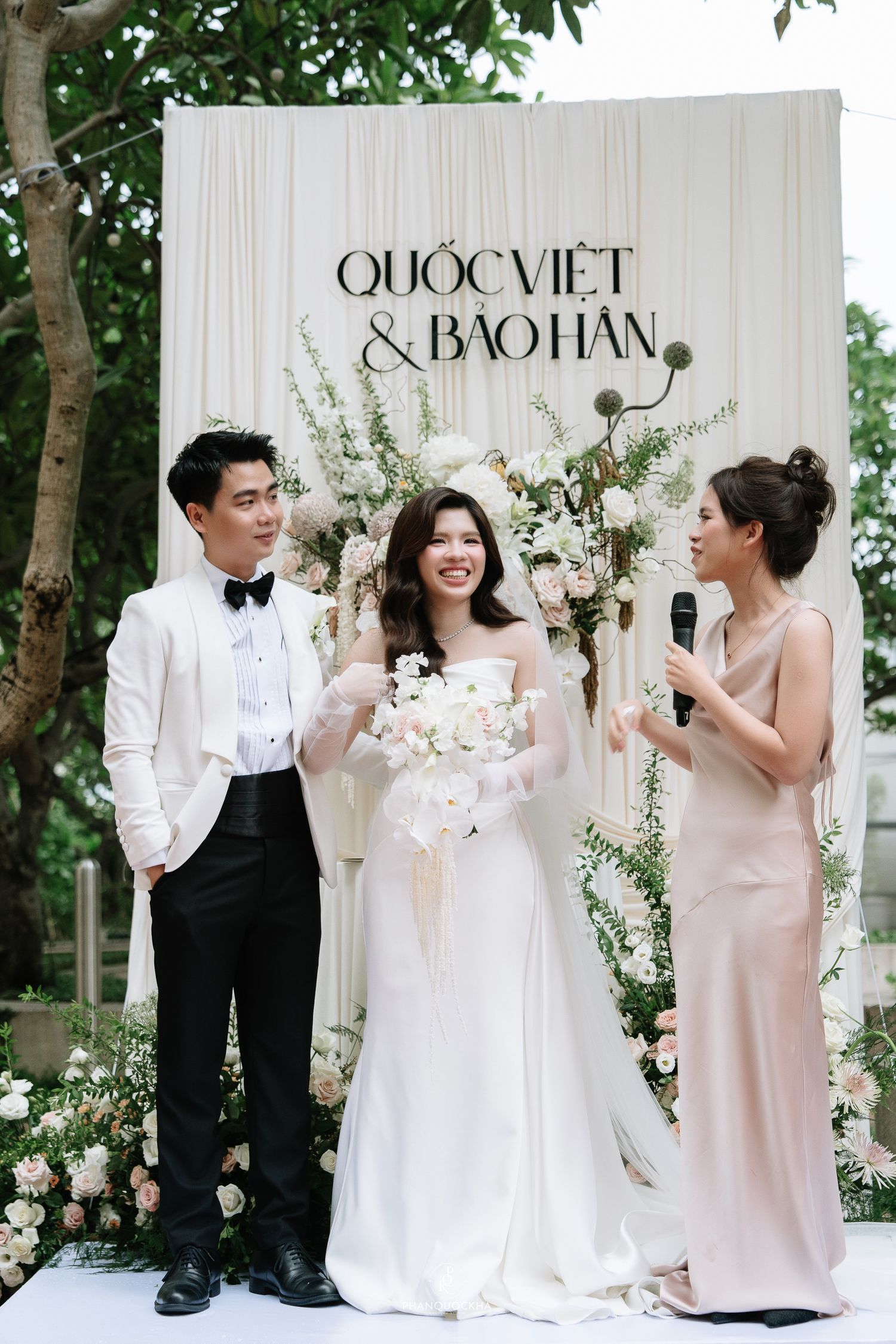 Bride and groom Quoc Viet and Bao Han smile with bridesmaid holding microphone at outdoor wedding ceremony.