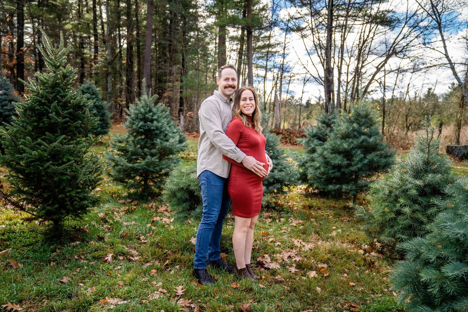 Couple selecting their perfect Christmas tree at a farm surrounded by evergreen trees and fallen autumn leaves.